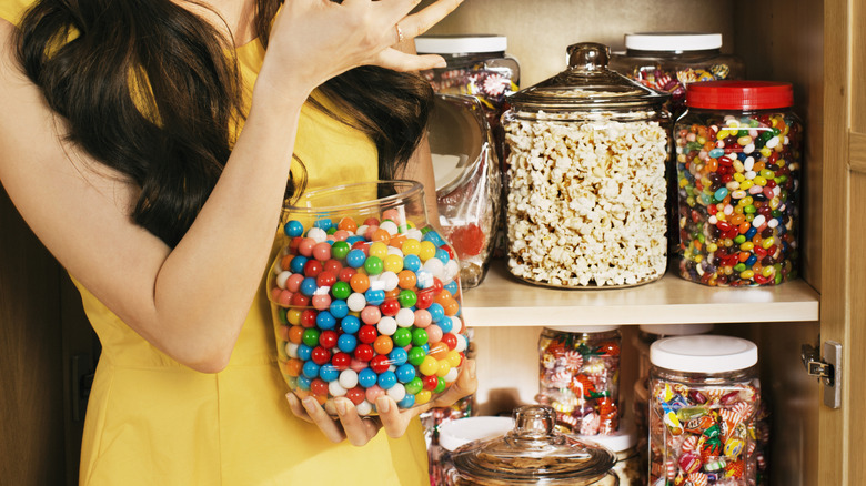 A person holding a huge jar of gumballs with more candy in the pantry