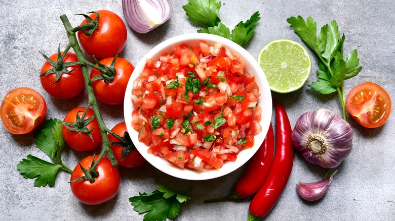 Bird's eye view of a bowl of salsa roja surrounded by ingredients (tomatoes, cilantro, onion, garlic, lime and chili peppers) on a grey surface