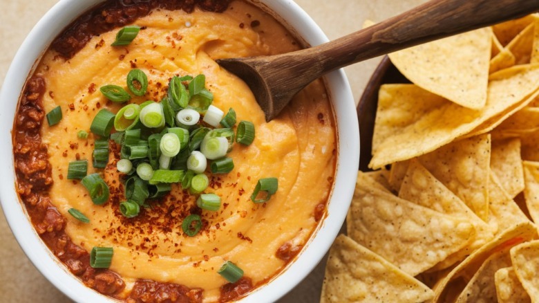 Chili dip topped with cheese sauce and green onions, served next to a bowl of tortilla chips