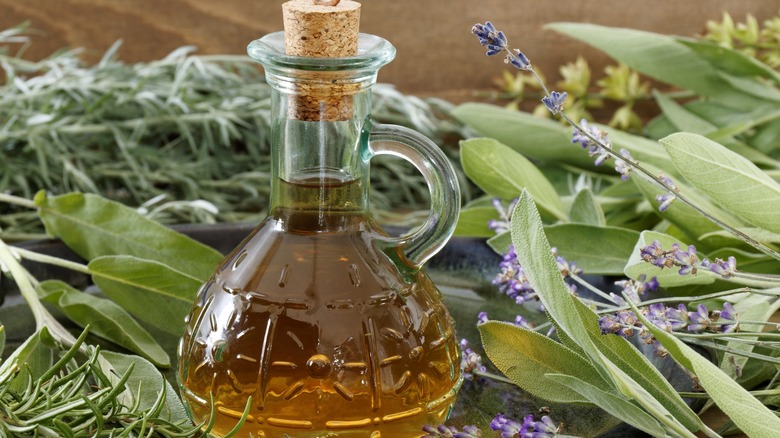 An ornate bottle of herb-infused vinegar, surrounded by sage and rosemary leaves, and lavender blossoms.