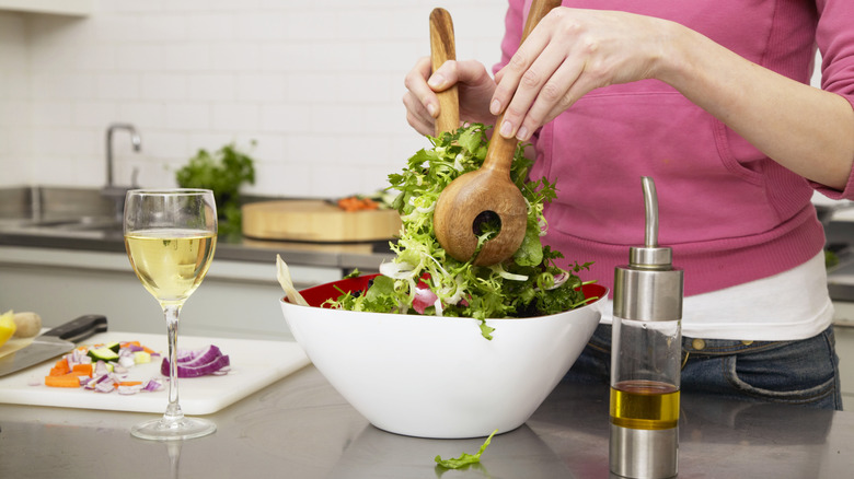 A woman tossing salad in a large bowl with a glass of white wine nearby