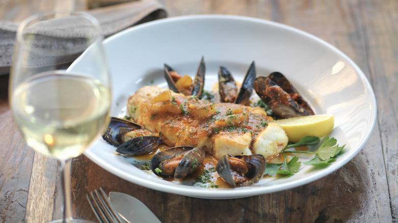 A seafood dish with fish and mussels behind a blurred glass of white wine