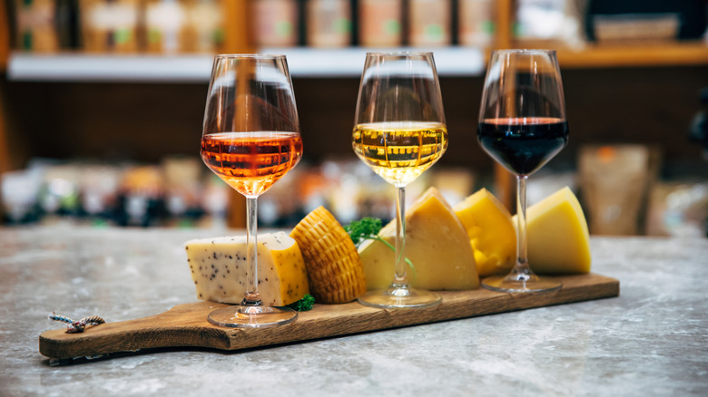 A cheese tray with different types of cheeses and three glasses of red, white, and rose wine