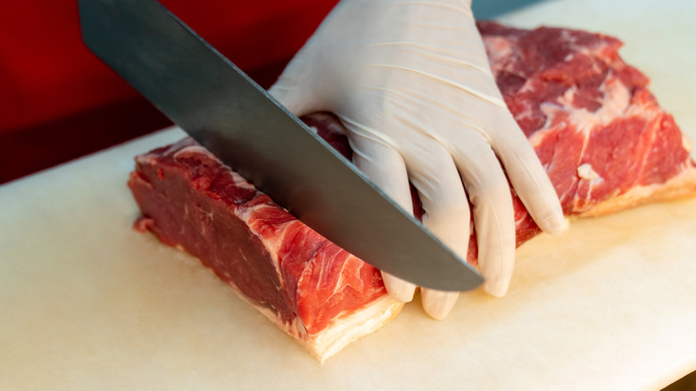 A butcher wearing white plastic gloves and slicing raw meat