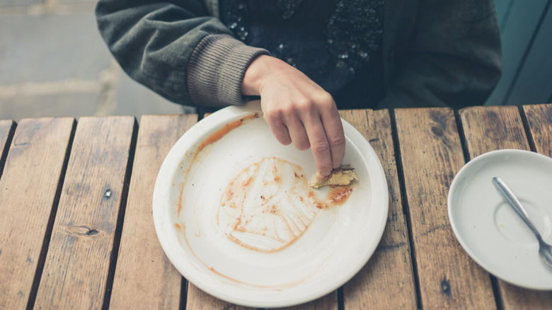 A person at a wooden table wiping up sauce from a white plate with a piece of bread