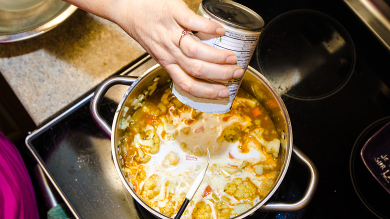 top view of creamy coconut milk being poured into pot of vegetable stew