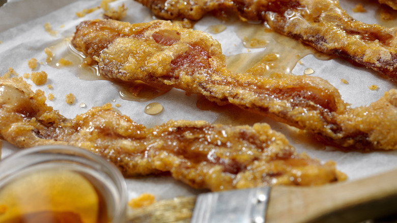 Candied bacon on a lined baking sheet with maple syrup on the side in a small glass bowl.