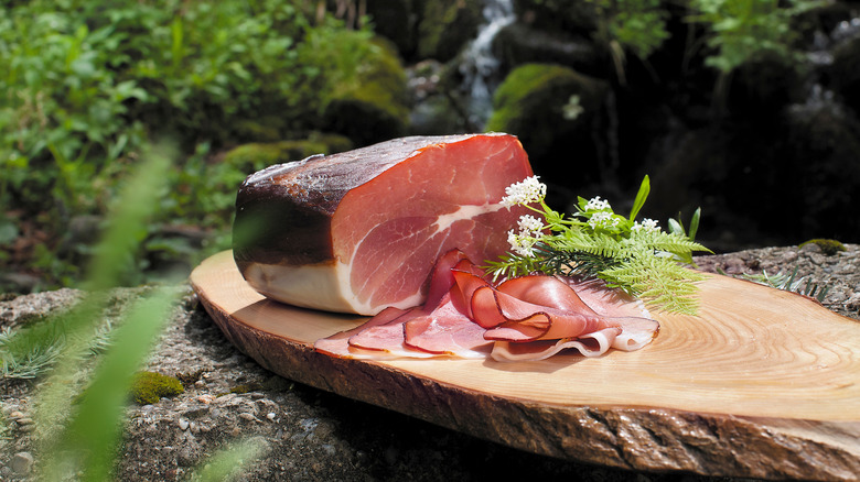 Black Forest ham sitting on a wood plank with some type of white flowers in an outdoor setting.