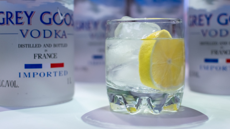 A glass of Grey Goose vodka with a lump of ice and a slice of lemon and vodka bottles in the background