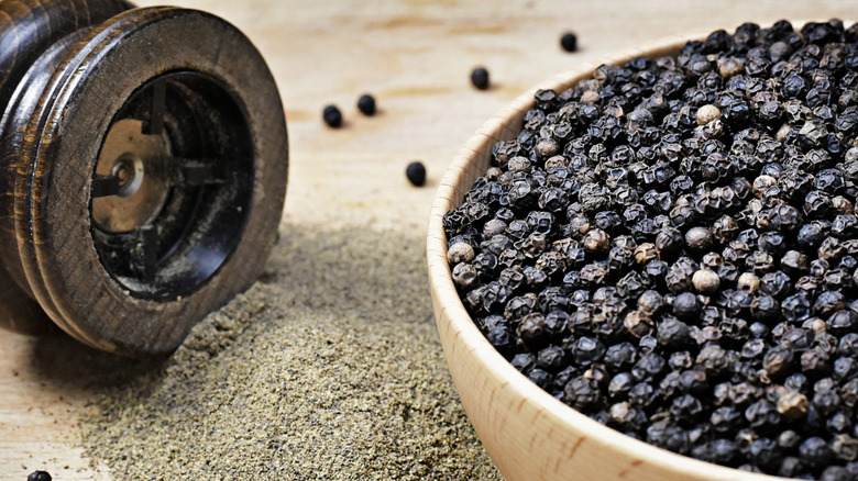 A bowl of black peppercorns beside a grinder and ground black pepper