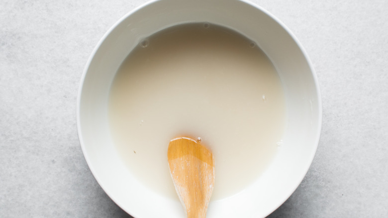 A white bowl of yeast and water sits on a white countertop with a wooden spoon in it