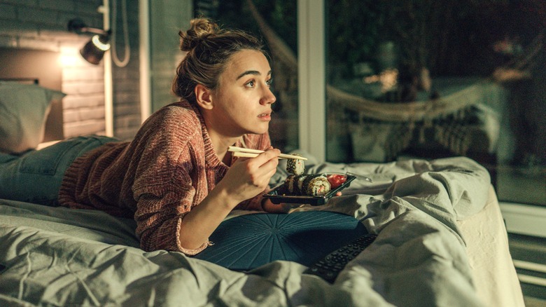 Woman lays in bed eating sushi while watching TV