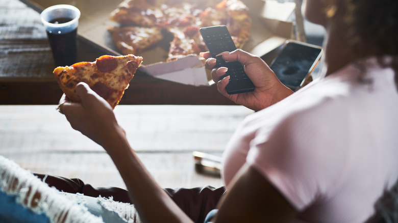 Woman sits on her sofa eating pizza and holding a TV remote