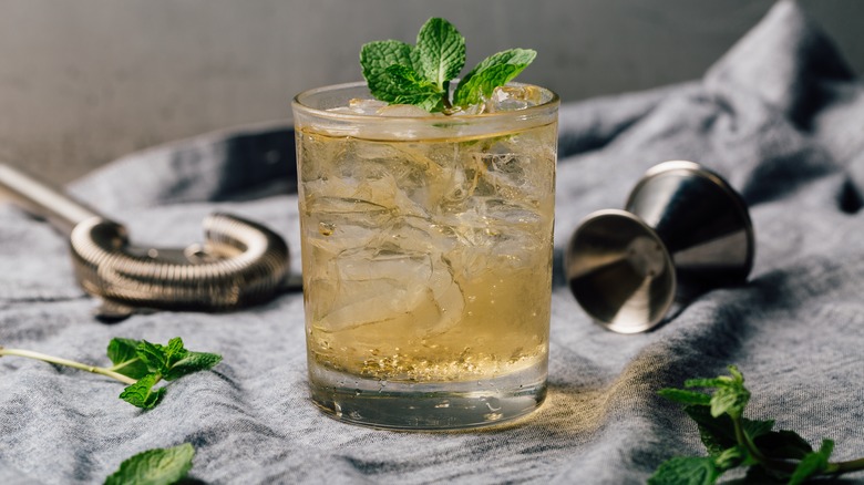 Whiskey smash against a blurred background with mint leave laying around the glass