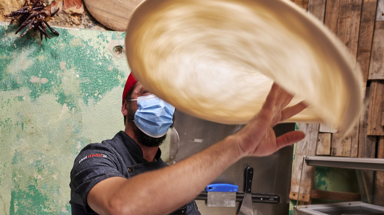 A chef with a mask on spinning pizza dough in his hand