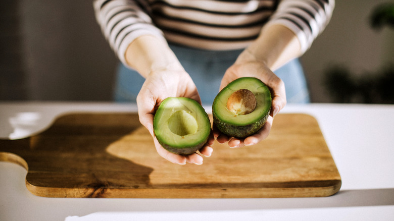 An individual holding an avocado split in half with a cutting board underneath