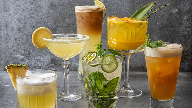 Six cocktails in various cups with different garnishes including lime, lemon, and pineapple