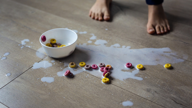 A bowl of cereal and spilled milk on hardwood floor.