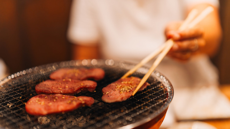 A person using cooking chopsticks to flip meat on grill.