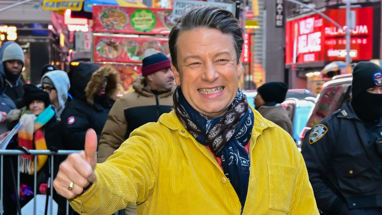 Jamie Oliver smiling and giving a thumbs up while walking in a city.