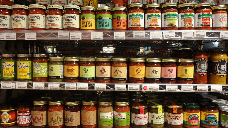 Many varieties of jarred salsa on grocery store shelf.