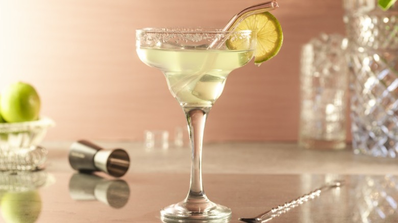 Margarita with lime wedge and glass straw, with bowl of limes and cocktail jigger in background.