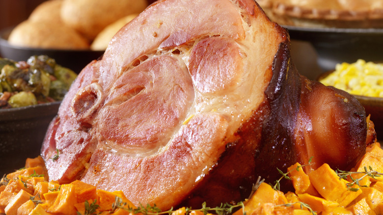 Cooked ham on a bed of sweet potatoes with other cooked sides such as corn nearby on a table