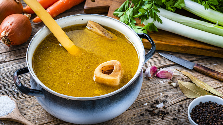 Bone broth in a ceramic pot on a wooden table surrounded by various vegetables like onions, carrots, and celery