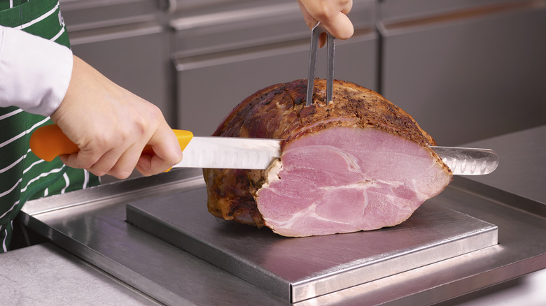 A person uses a carving knife to slice a ham on a cutting board