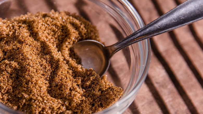 A glass bowl of dark brown sugar with a metal spoon.