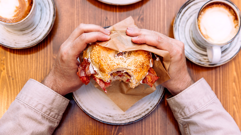 Pair of hands holding a breakfast sandwich with bacon, next to two cups of coffee.