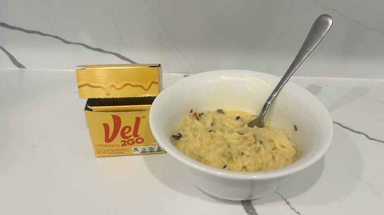 A bowl of cheesy ramen noodles is shown next to Vel2Go packets