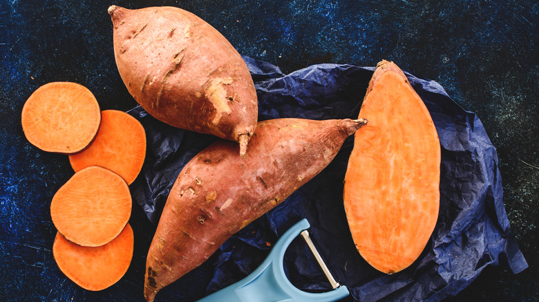 Whole and sliced sweet potatoes on blue background with a peeler