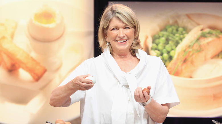 Martha Stewart laughs in a demo kitchen holding eggs in a white shirt.