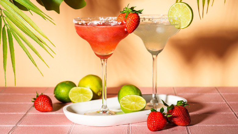 Two margaritas, one strawberry and one lime, accompanied by limes and strawberries