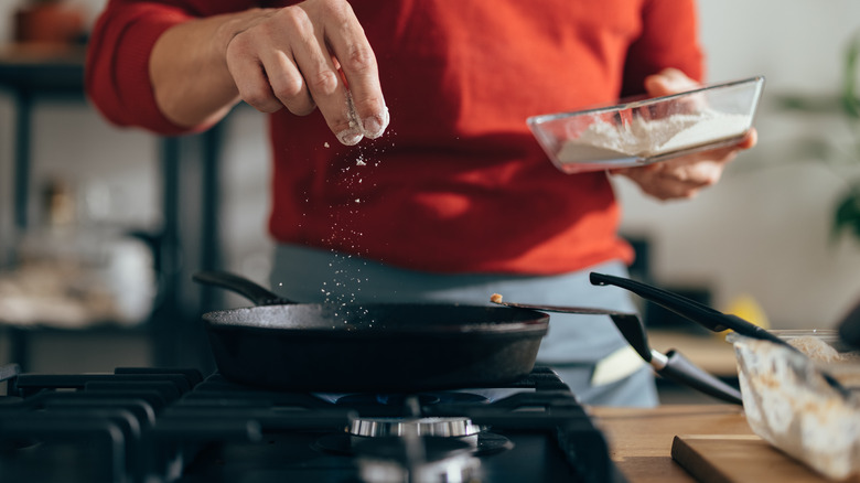 A man in a red shirt adding salt from a bowl to a pan on the stove.