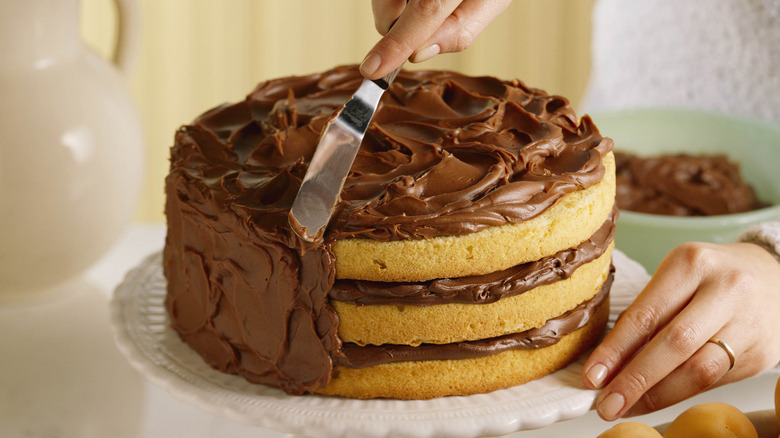 Hands icing a three-tier yellow cake with chocolate frosting