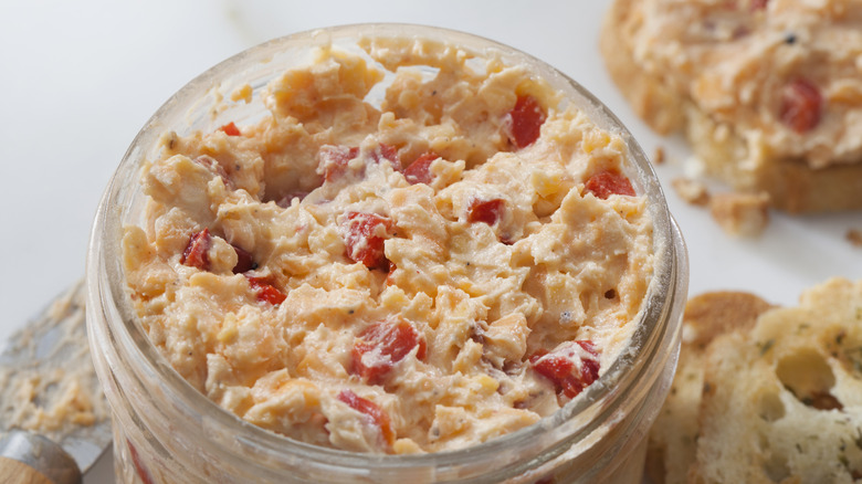 Glass jar full of pimento cheese on a light grey surface with toast and a knife for serving.