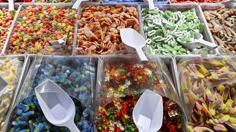 A wide selection of gummy candies are seen in metal containers with plastic scoops and bags