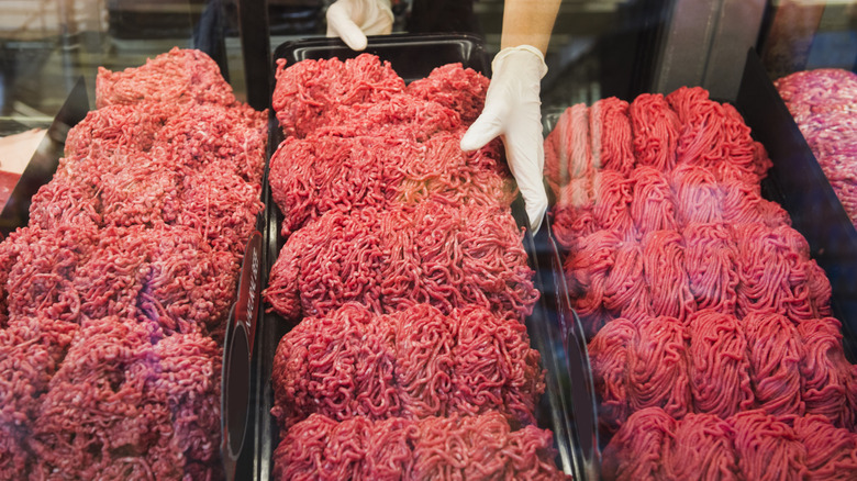 Three large black trays filled with ground beef behind glass with a glove-covered hand adjusting one.