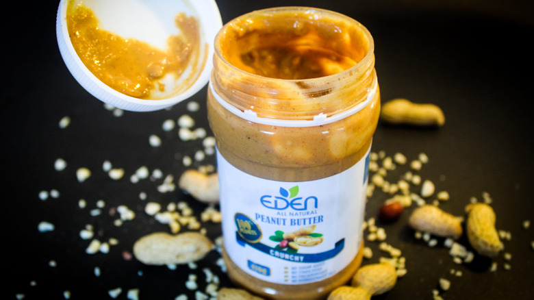 Jar of crunchy peanut butter with the lid open, surrounded by crushed and whole peanuts