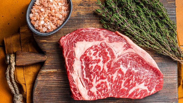 A raw piece of well-marbled steak on a wooden board, with thyme and kosher salt nearby.