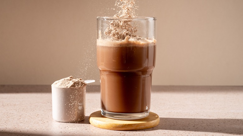 protein powder falling into a glass full of protein shake, with a scoop full of protein powder beside the glass