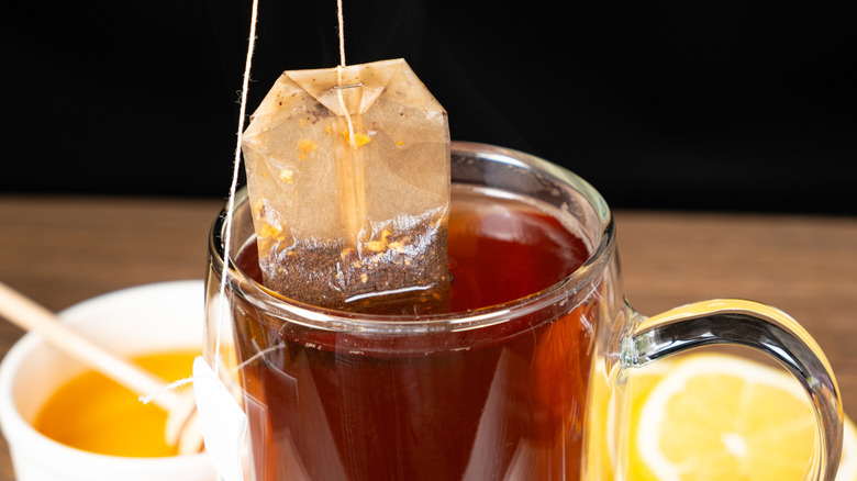 Tea bag being steeped in a clear cup of water behind which are a a white container with honey and a lemon cut in half.