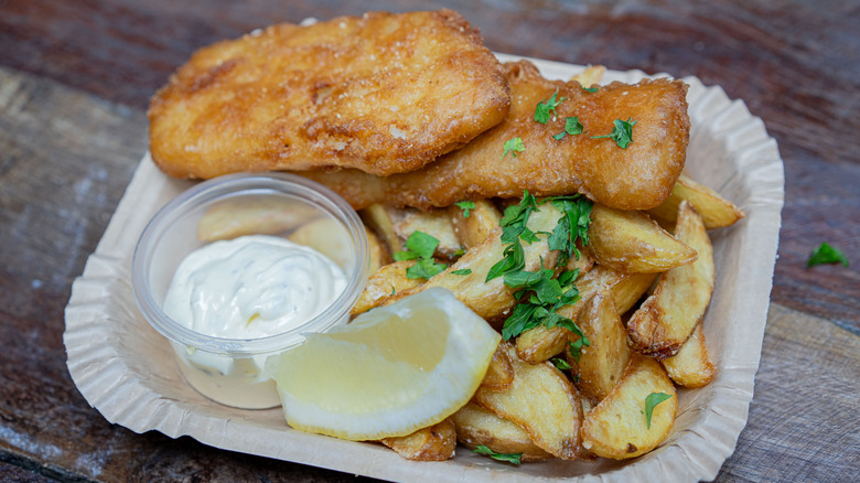 A rectangular paper plate with fish and chips, tartar sauce and a lemon slice on it.