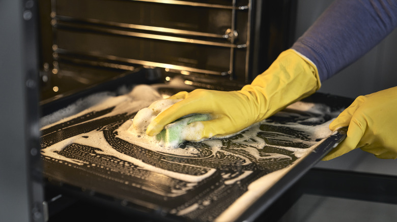 A close up of hands in yellow gloves cleaning an oven door with a green sponge