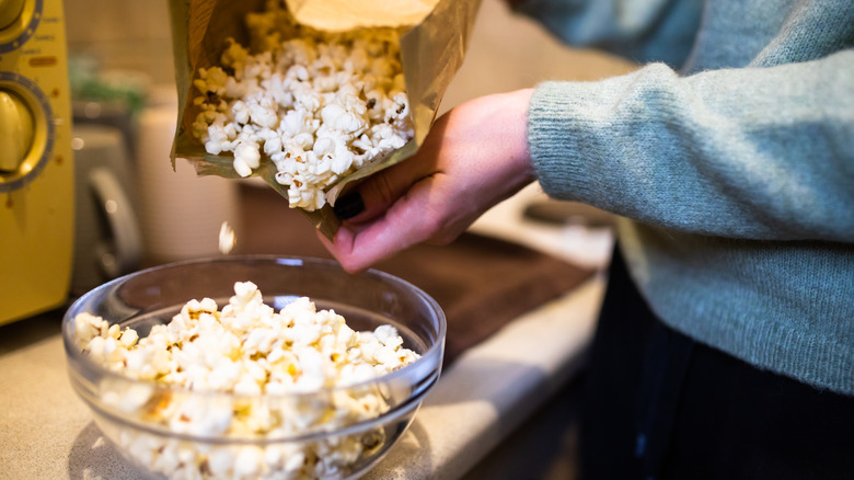 A person pouring a microwaved bag of popcorn into a glass bowl on a kitchen counter.