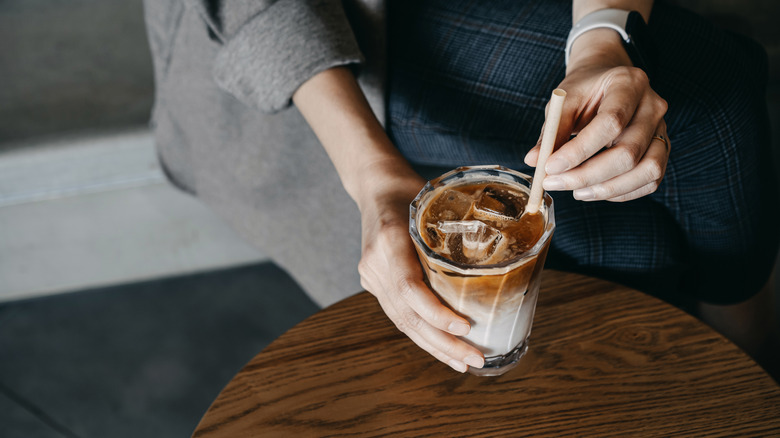 Woman sitting at a table stirring an iced coffee drink with a straw