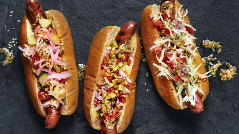 Three hot dogs in buns with pickles, tomatoes, onions and more on top