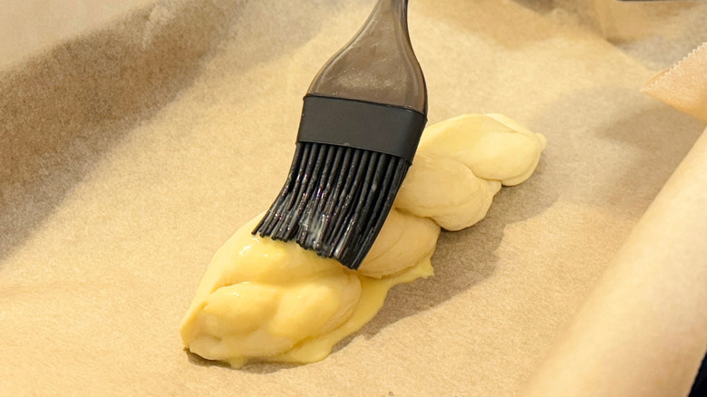 A person applies egg wash to pastry sitting on brown paper in a pan.